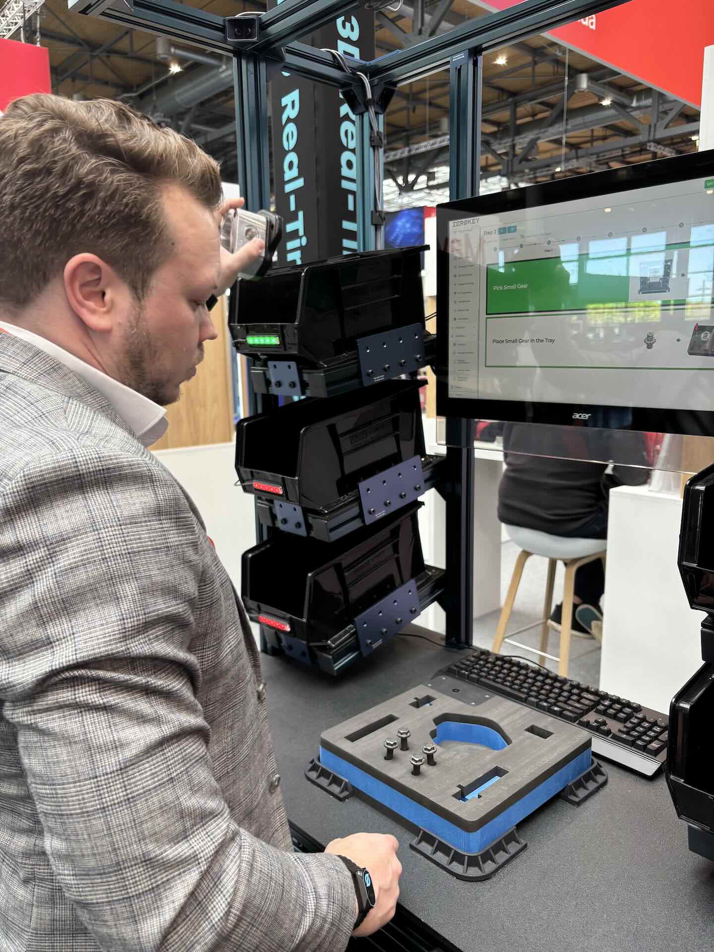 A man wearing a gray plaid jacket interacts with a computer setup at a trade show. The screen displays software, and black storage bins are stacked on either side of a keyboard and device.