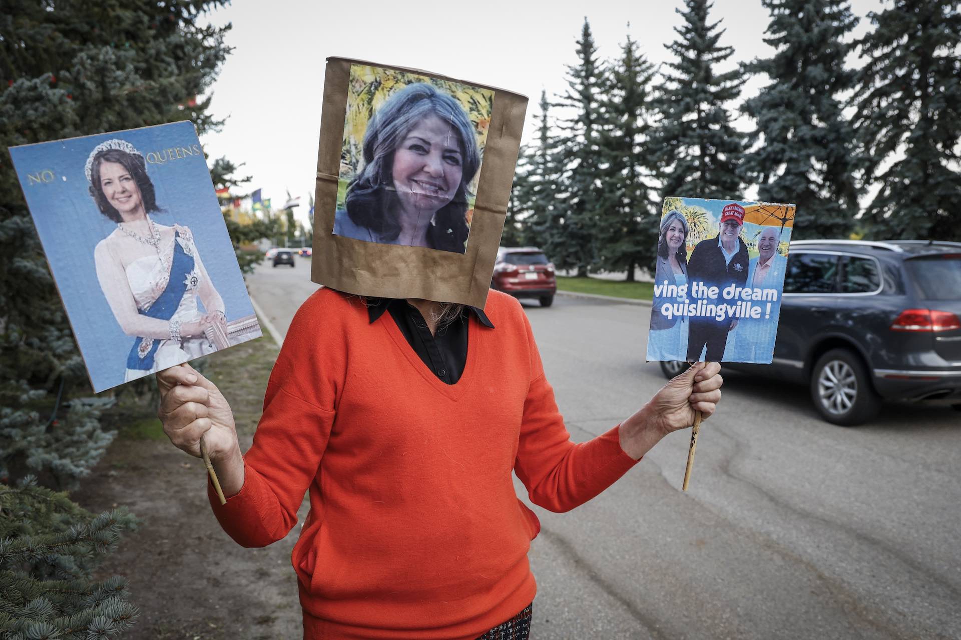 A shot of a female protester by a roadside wearing a paper bag over her head bearing an image of Danielle Smith's face. The demonstrator has a sign in each hand—one showing Smith mocked up as a queen, the other showing Smith with Donald Trump and Kevin O'Leary.