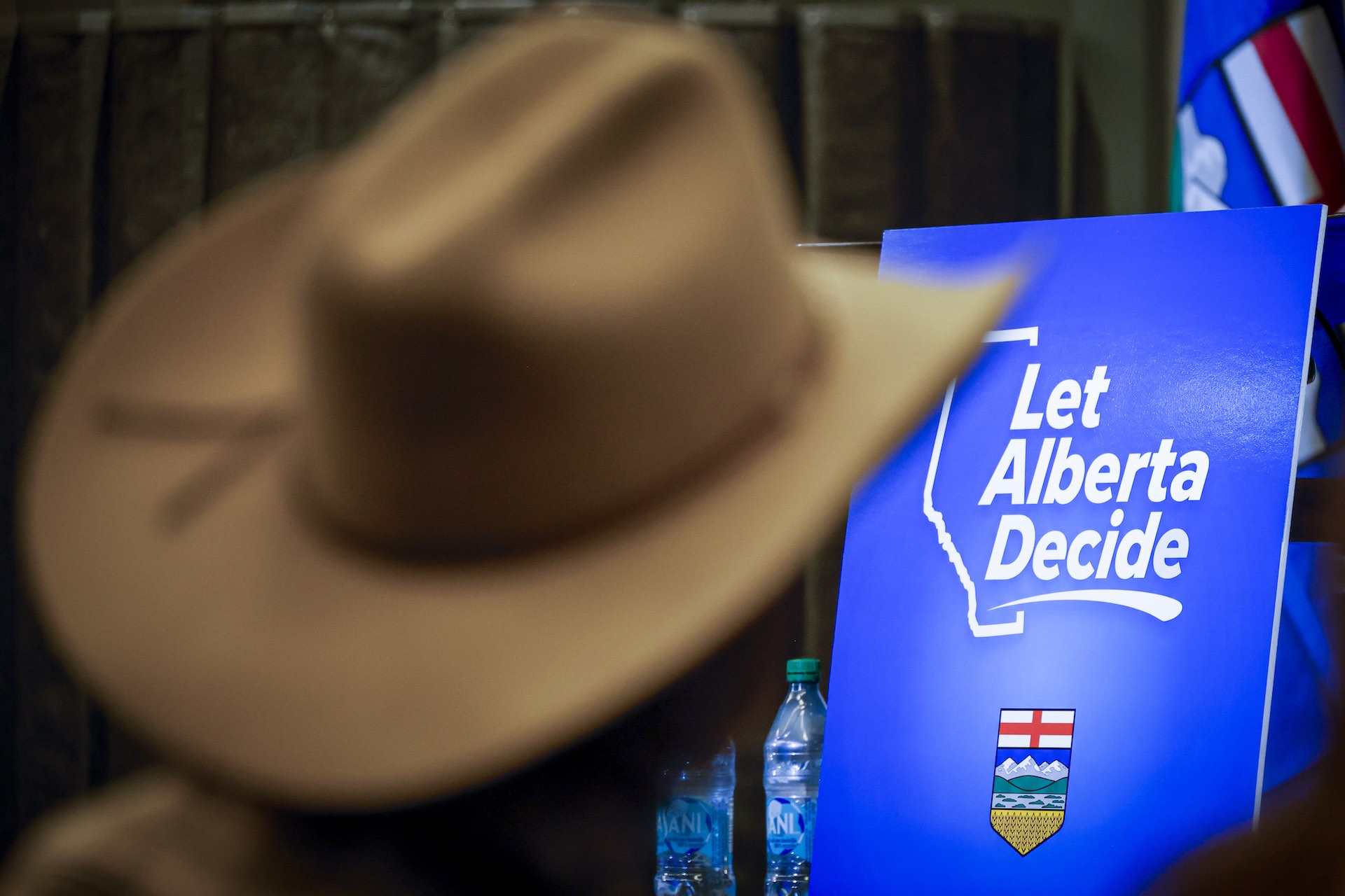 A shot of a placard on a table reading "Let Alberta Decide." There is a person out of focus in the foreground wearing a cowboy hat.