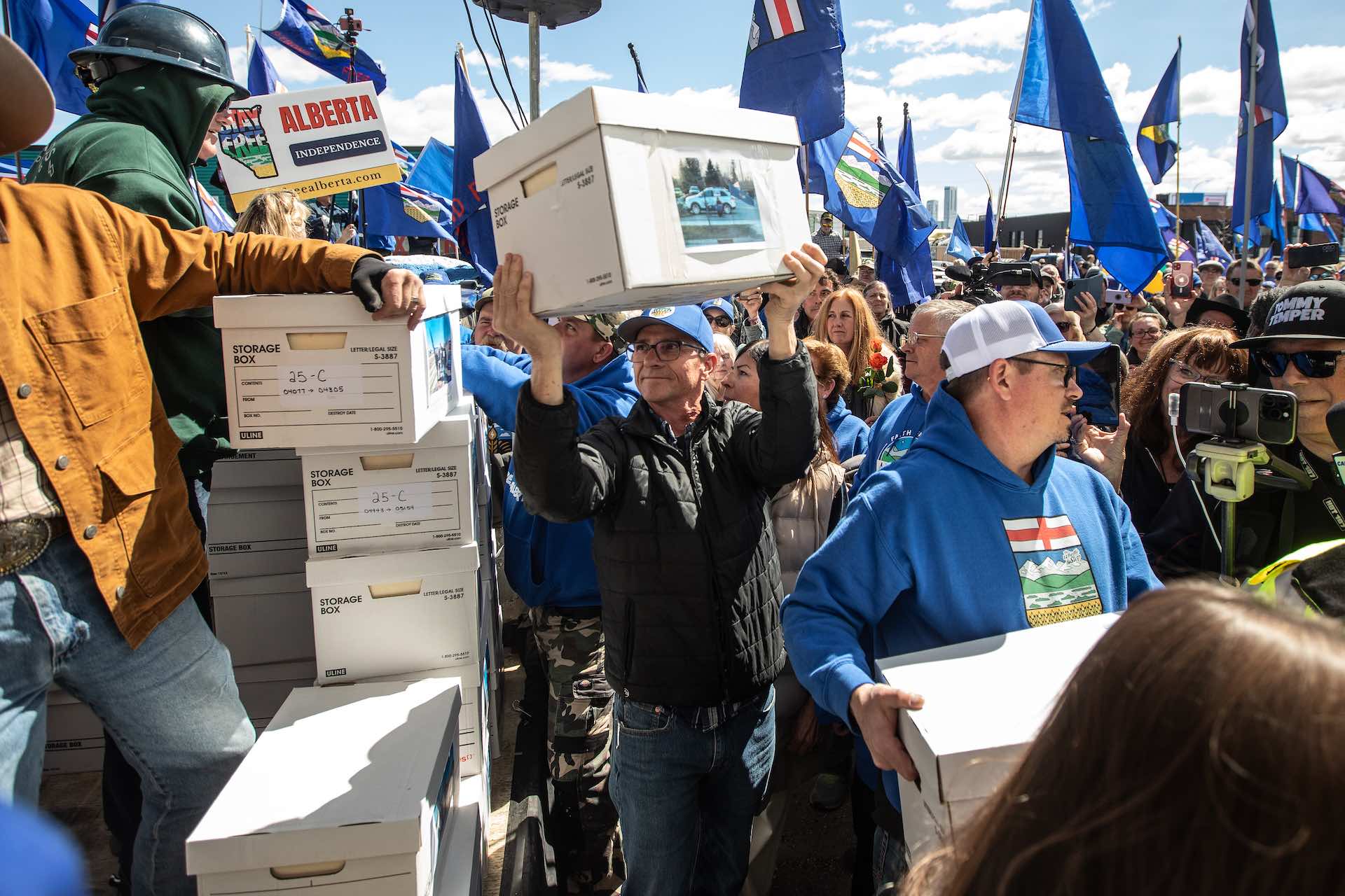 A shot of a crowd bristling with Alberta provincial flags. There is a man in the foreground hoisting a banker's box toward a stack of similar boxes.