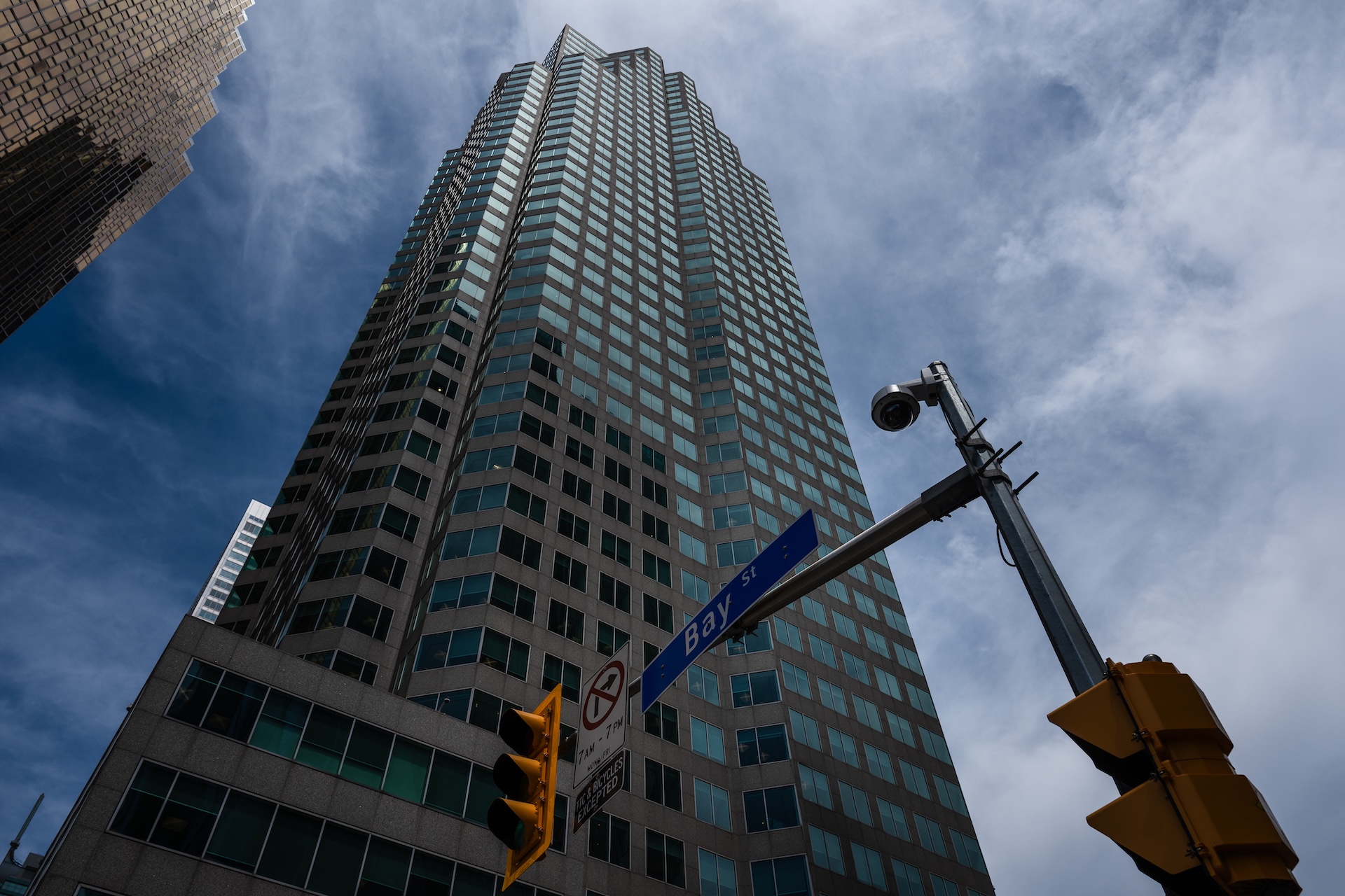 A skyscraper on Bay Street in Toronto, viewed from street level looking up, with a traffic light and street sign in the foreground against a blue sky with clouds.
