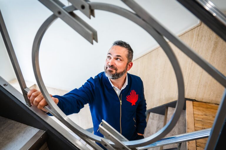 A photo of Daniel Sax shot through a circular piece of ironwork on a stairway balustrade. He's looking off-camera, and is wearing a dark blue jacket bearing his company's logo.