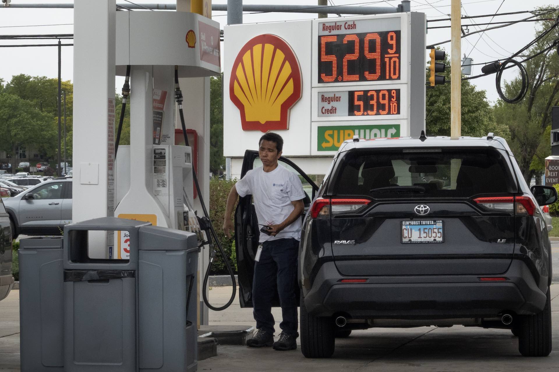 A shot of a man getting out of his car beside a set of pumps to fuel up. A giant Shell sign displays the price of gas at just below US$5.30 per gallon.