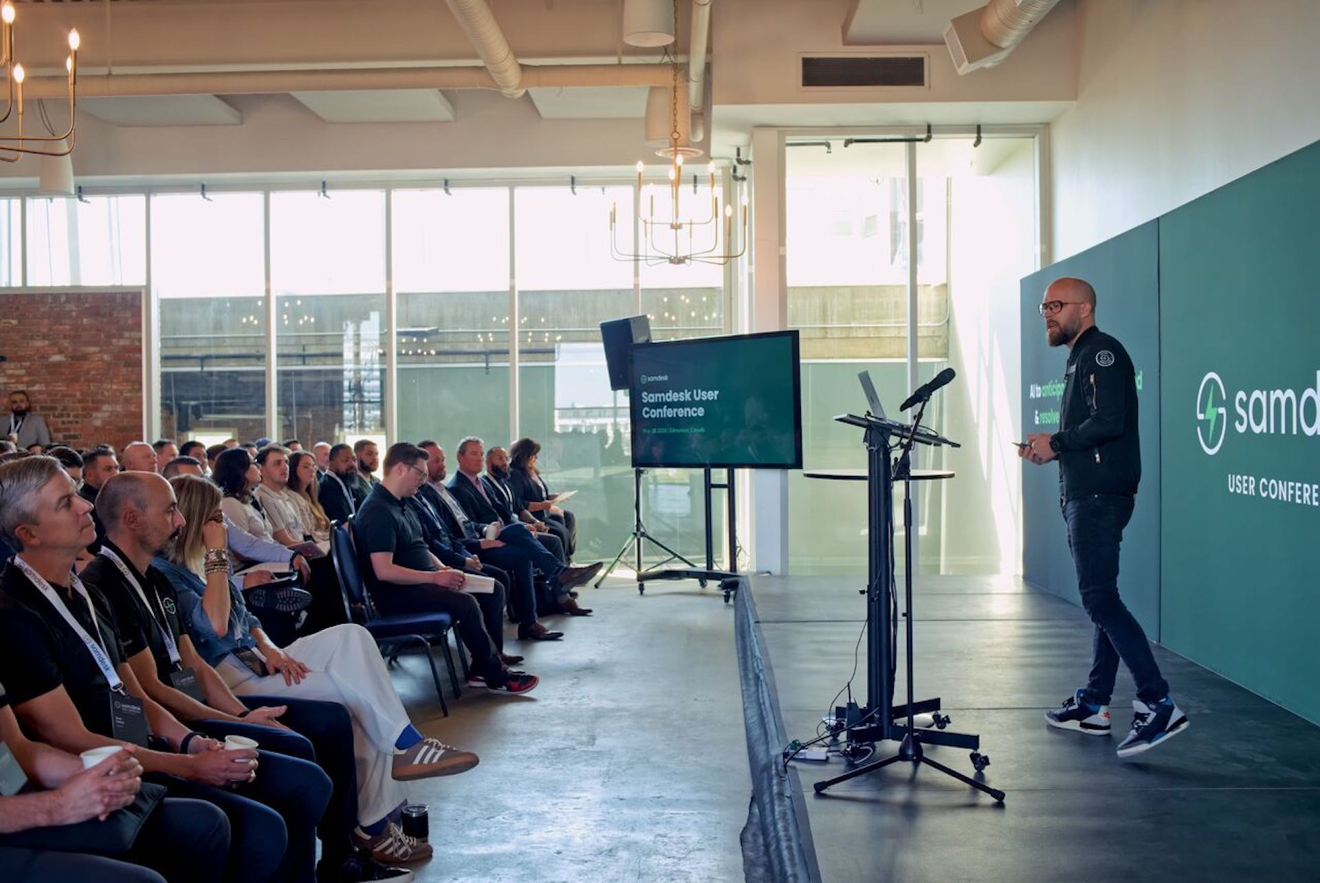 A side shot of James Neufeld speaking onstage before an audience. He's wearing black clothing and high-top running shoes. Samdesk's logo appears on a backdrop, and on a video monitor beside the stage.