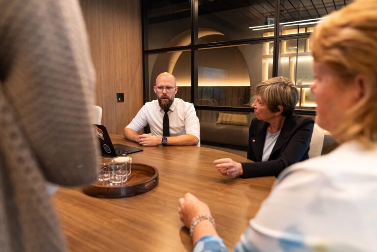 A head-on shot of James Neufeld seated with others at a round table in a meeting room. Eleanor Olszewski is seated to his left. There's a laptop open in front of Neufeld.