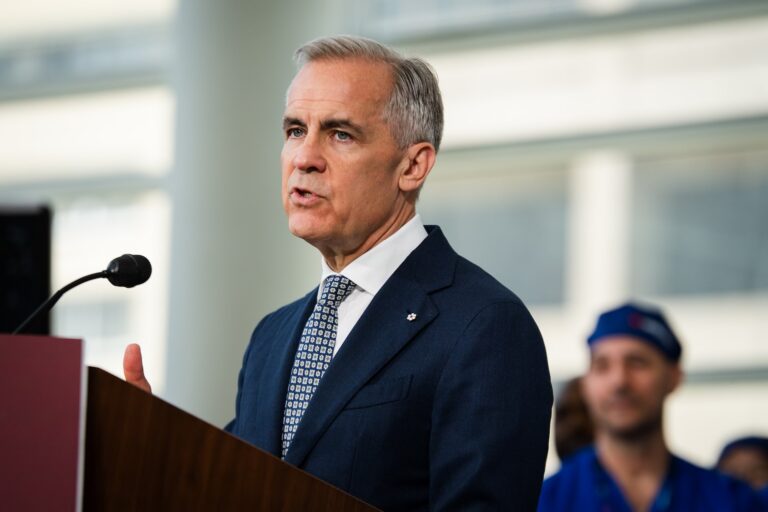 An image of Prime Minister Mark Carney speaking at a podium with a microphone. He is wearing a dark suit and tie. A person wearing scrubs is visible in the background.