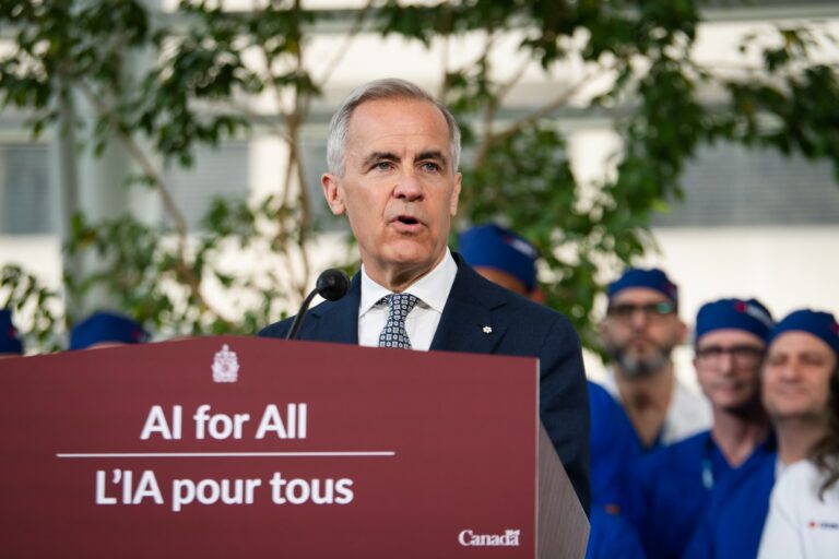 An image of Mark Carney standing in front of a red podium with the words "AI for All / L'IA pour tous." He is wearing a suit and tie. In the background, people wearing scrubs and white coats are visible.