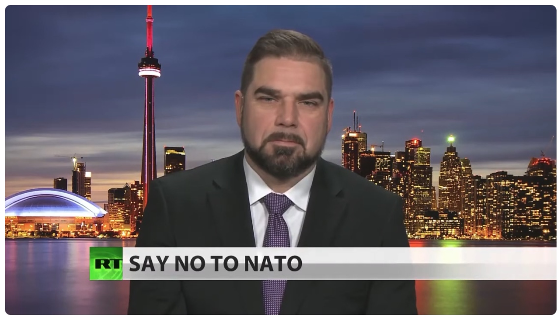 A screencapture of an RT broadcast with the words "Say no to NATO" across the screen. A man wearing a dark suit and tie looks directly into the camera, with a serious expression. The city of Toronto skyline is in the background.