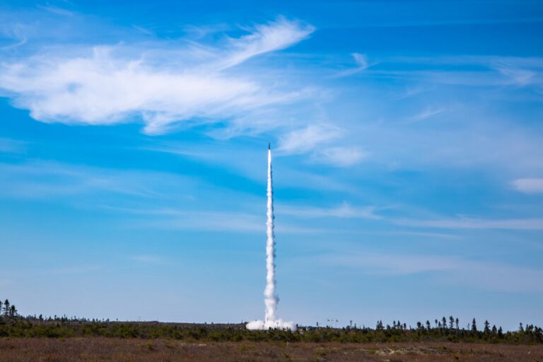 A shot across an expanse of low forest of a rocket launching into blue skies.