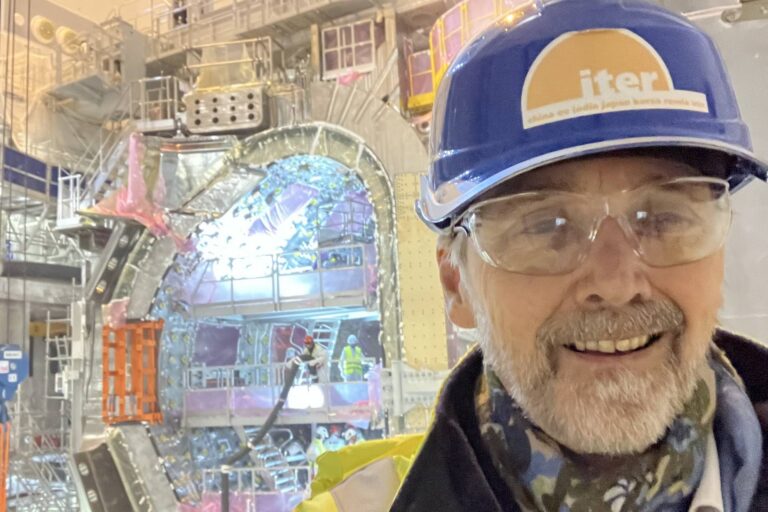 A selfie taken by Spencer Pitcher inside a nuclear fusion facility. He is wearing a blue hardhat with the ITER logo on it, and is standing in front of a cavernous chamber full of fusion reactor equipment.