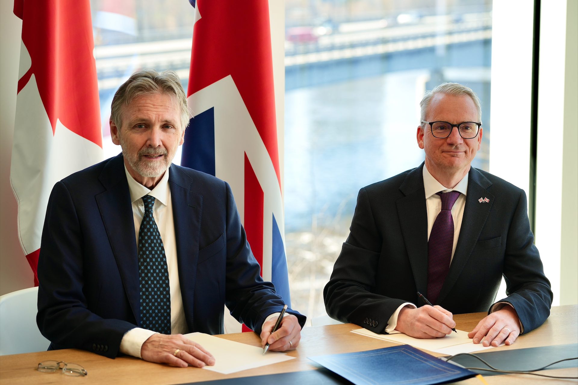 Two men in suits sit side-by-side at a table signing papers. There are U.K. and Canadian flags behind them.
