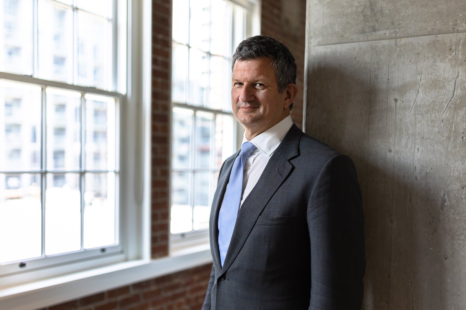 A portrait of Steve Hasker in a suit and light blue tie, standing against an exposed concrete wall, with high windows to his right.