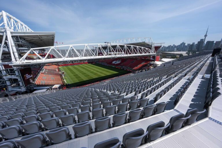 The image shows the inside of Toronto Stadium on a sunny day. The rows of seats are empty; an empty green field is visible.