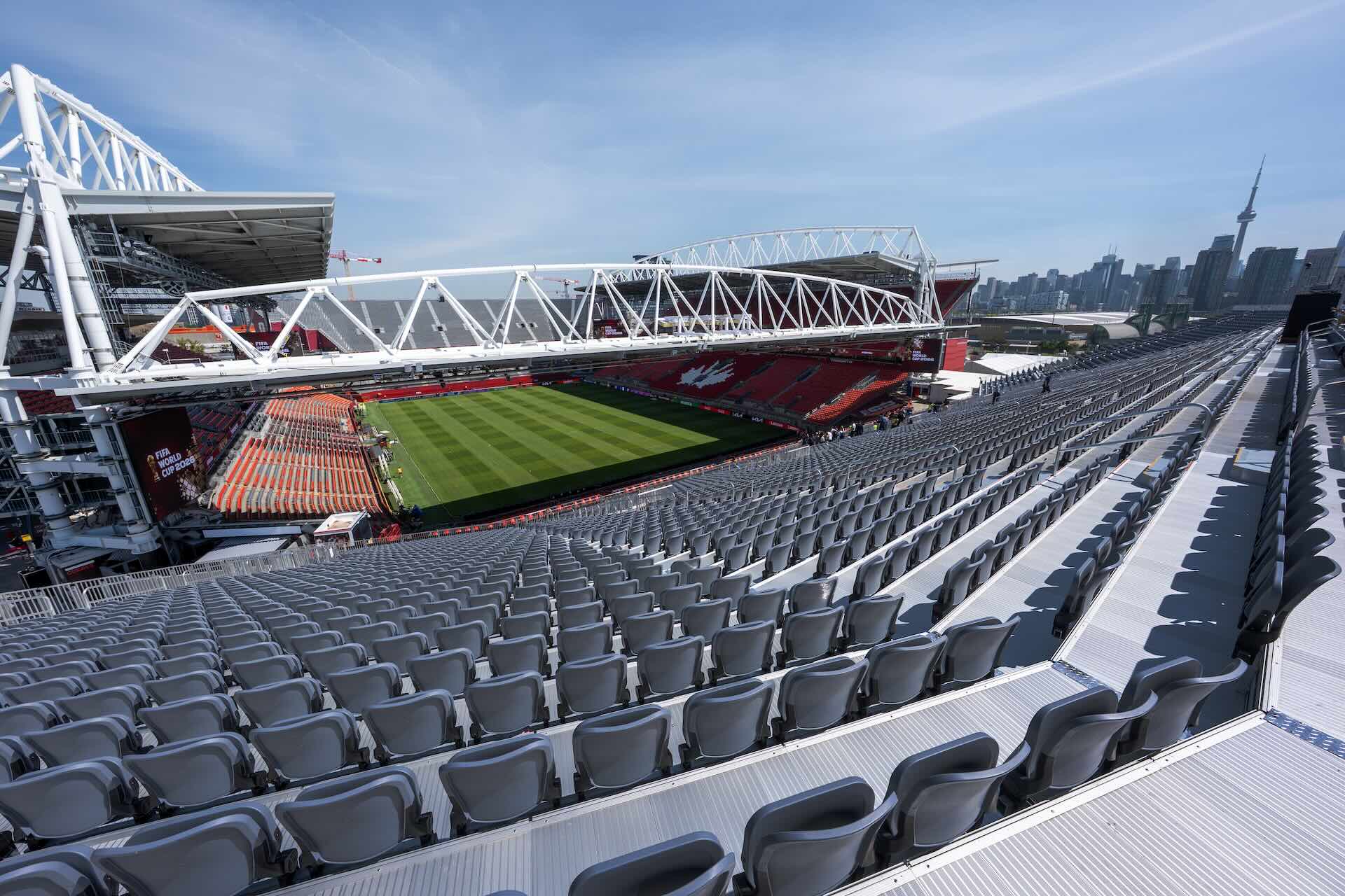 The image shows the inside of Toronto Stadium on a sunny day. The rows of seats are empty; an empty green field is visible.