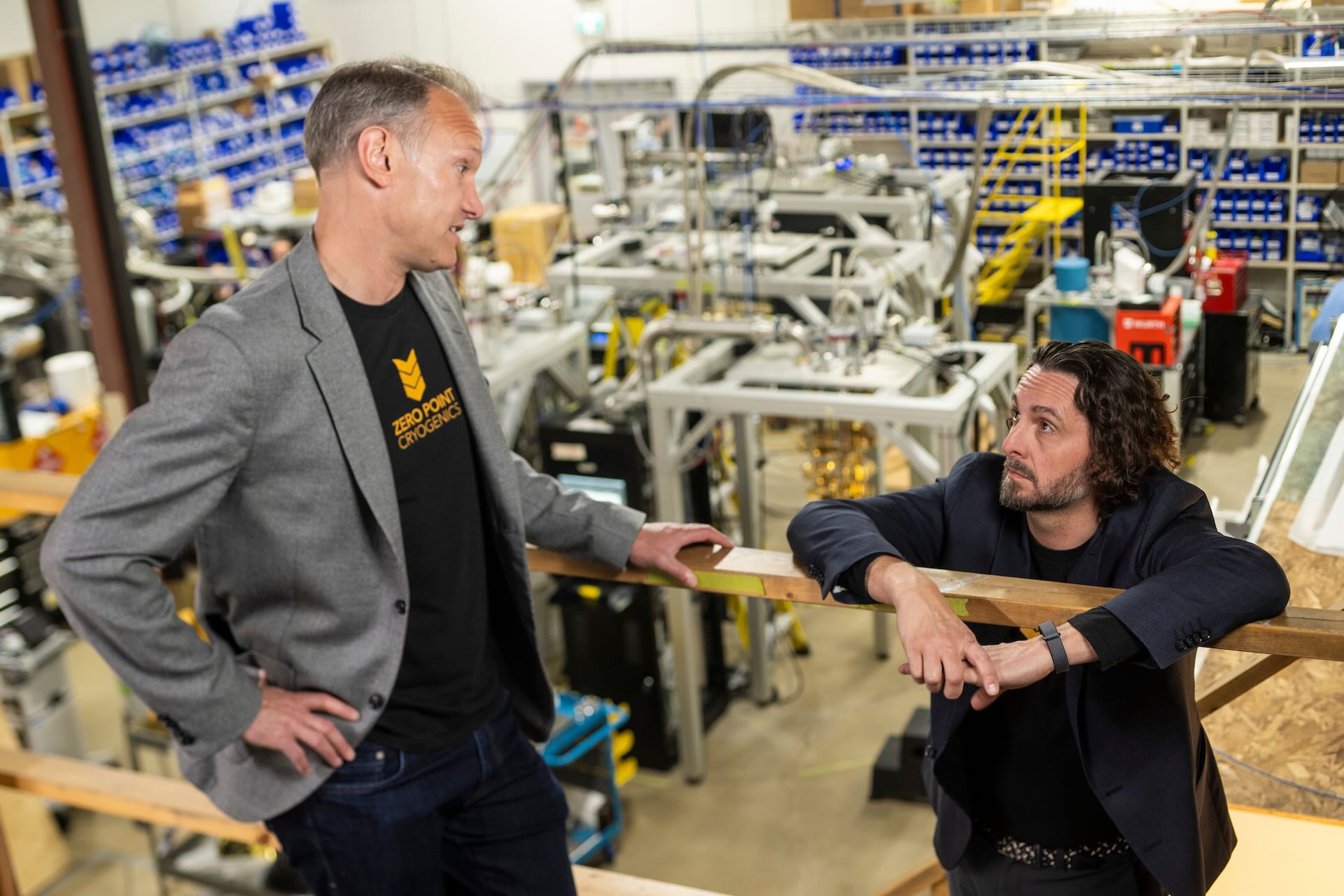 A shot of Christopher Cassin and John P. Davis in discussion; they’re on a catwalk above a shop floor that is crowded with equipment.