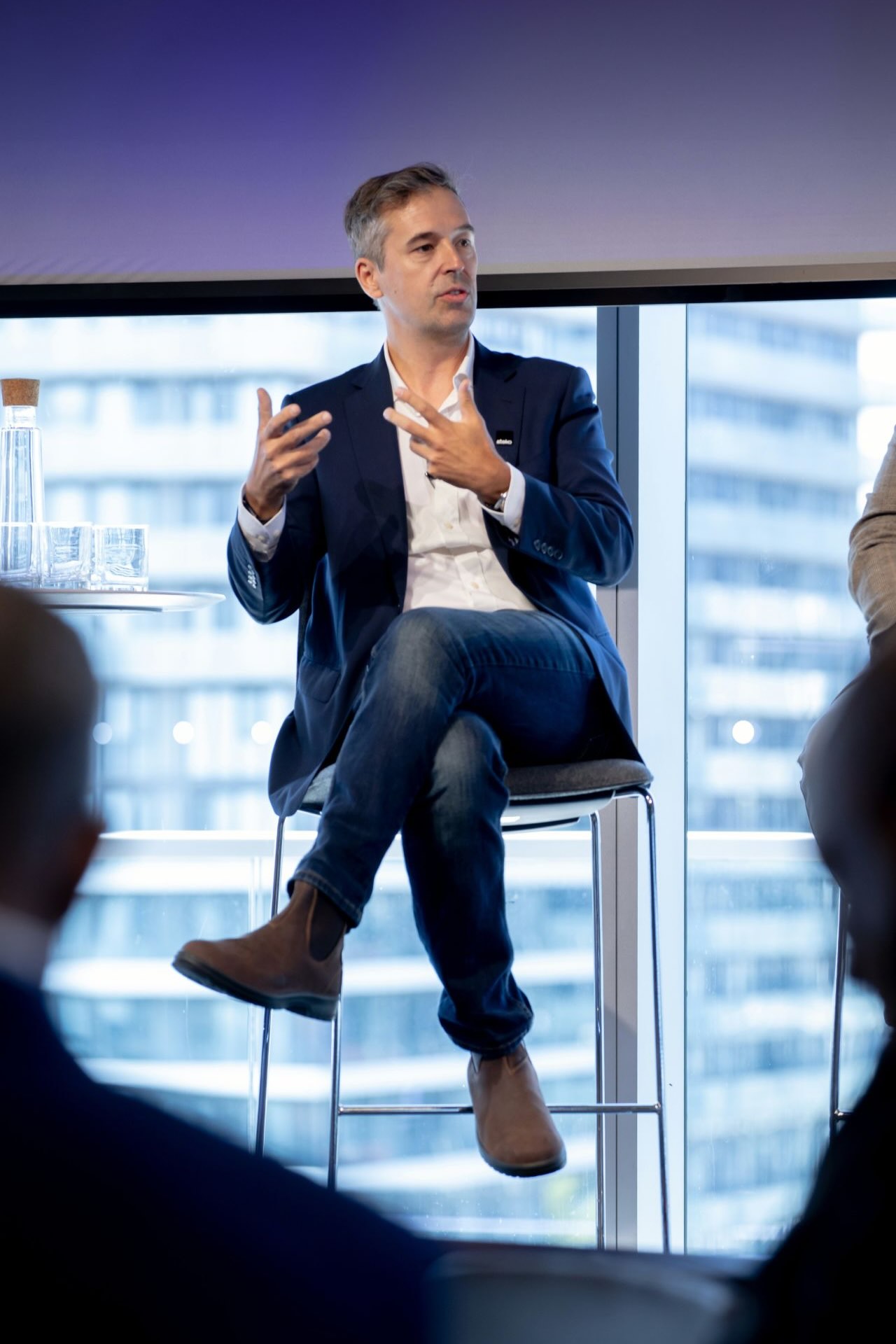 A man in a blazer and jeans sits on a high chair, gesturing while speaking during a panel discussion. Cityscape visible through a large window behind him.