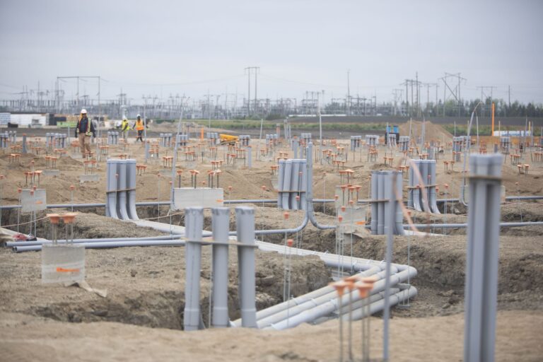 Workers at a large construction site with rows of grey pipes and electrical infrastructure in the background.