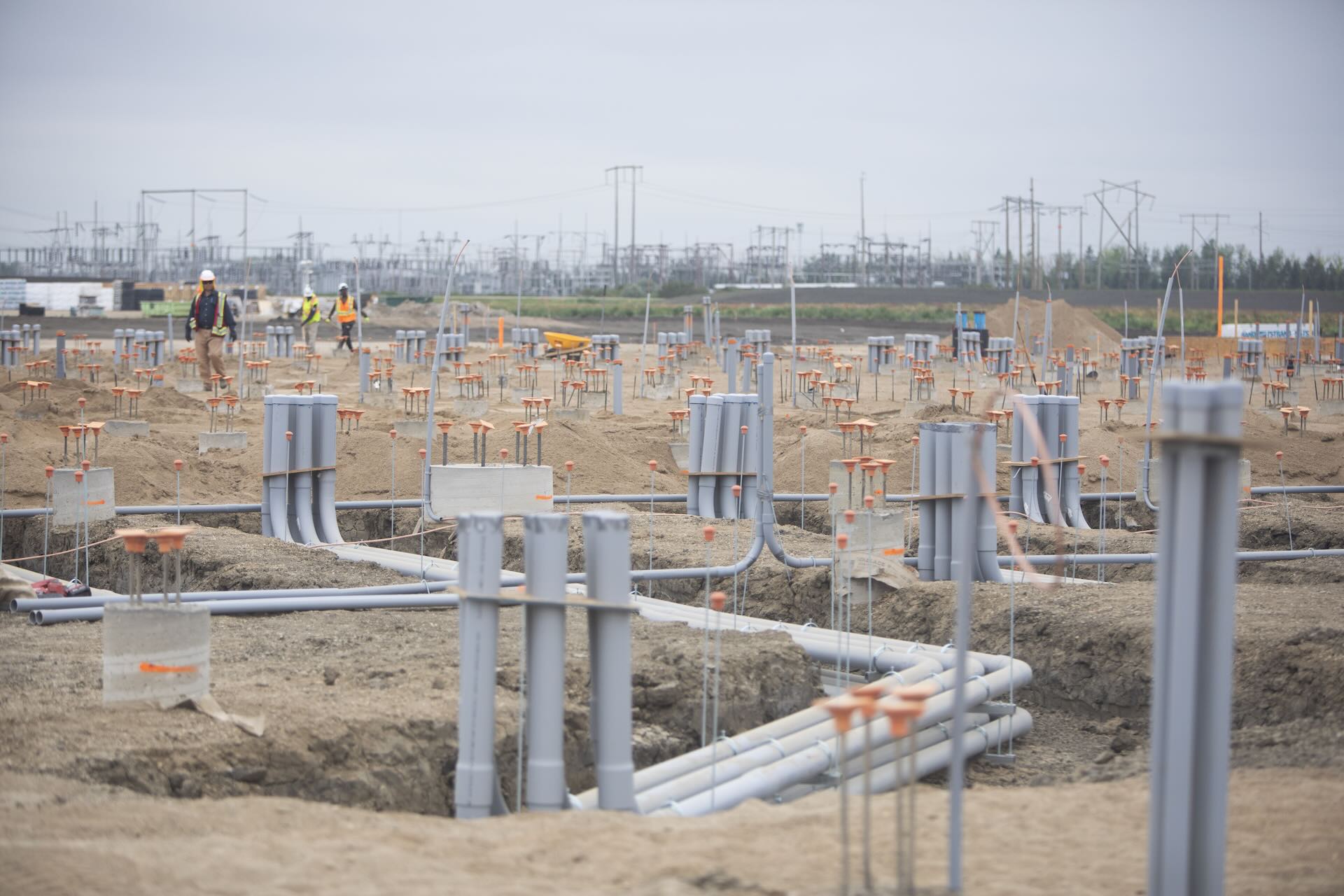 Workers at a large construction site with rows of grey pipes and electrical infrastructure in the background.