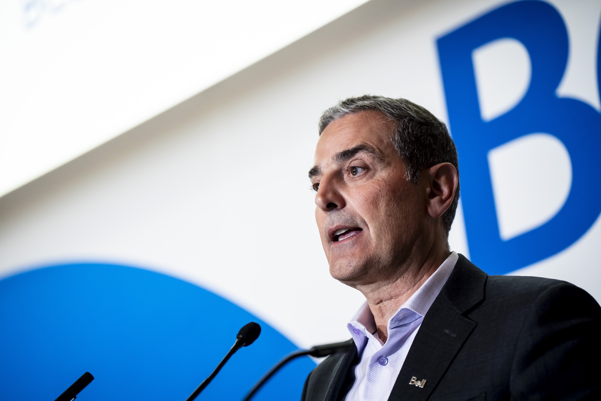 Bell CEO Mirko Bibic speaking at a podium in front of a microphone. He wears a dark suit jacket over a light blue shirt, in front of a blue and white backdrop with a partially visible letter “B” logo.