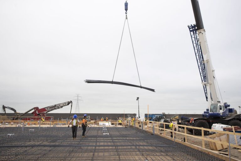 Construction workers walk across a deck as a crane lifts a large bundle of steel rebar into place at a construction site.