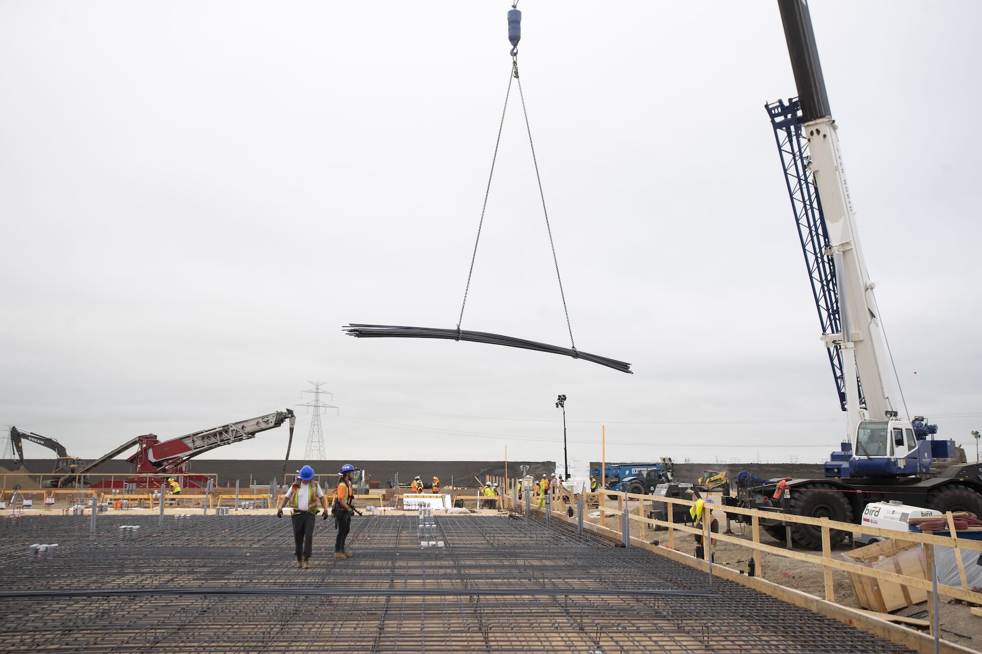 Construction workers walk across a deck as a crane lifts a large bundle of steel rebar into place at a construction site.