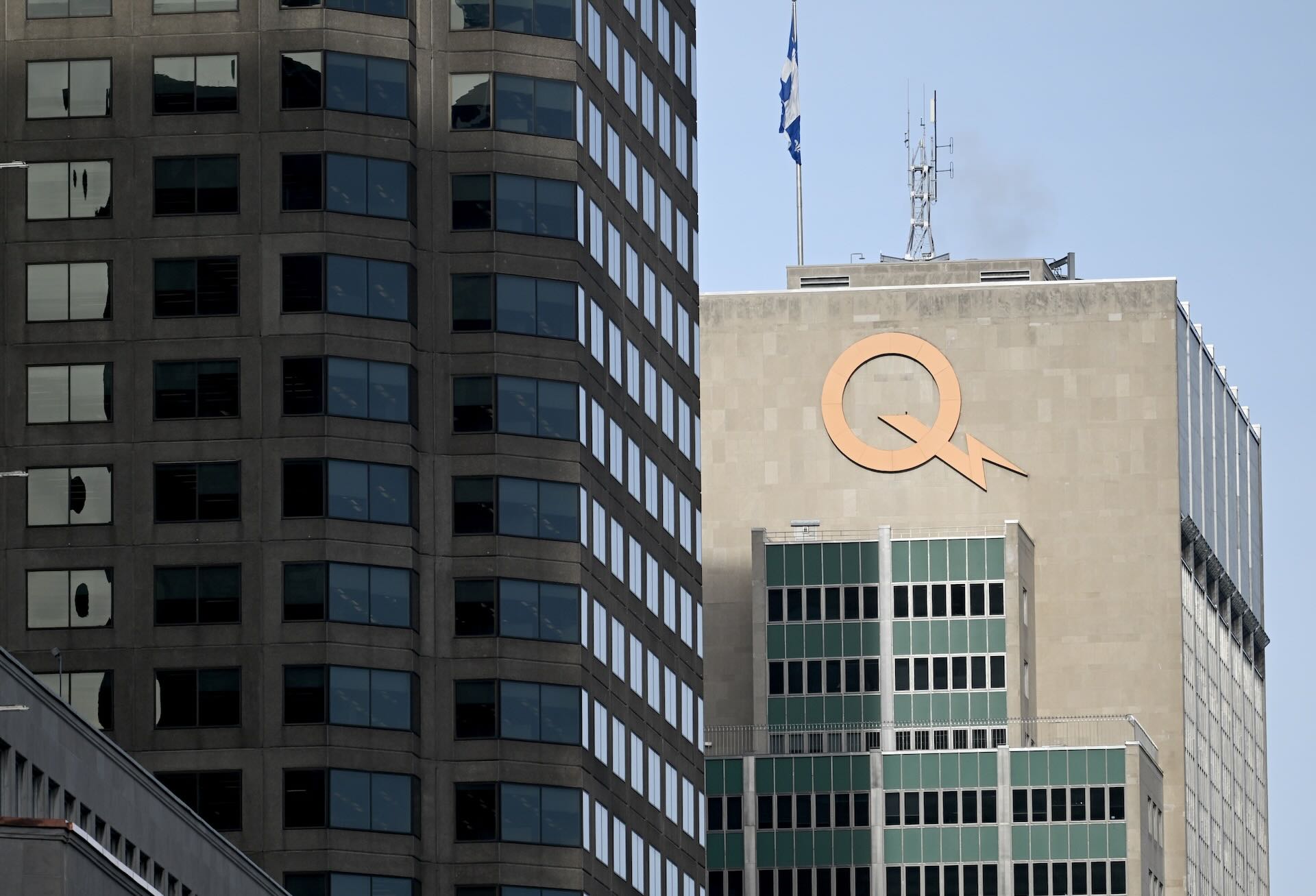 A cityscape featuring two tall buildings; the right one has a large orange "Q" logo and a Quebec flag atop. The sky is clear and blue.