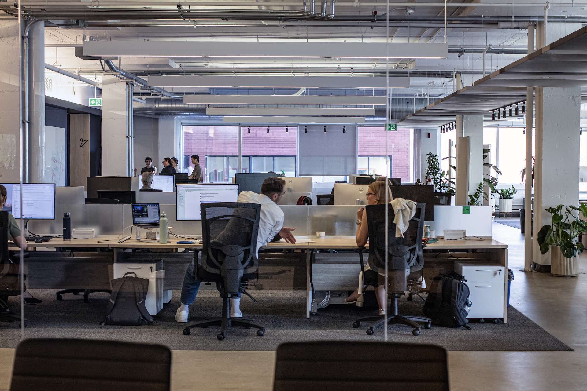 A wide shot of LawZero's industrial-chic, open-concept office. There are two people at desks in the foreground and a small cluster of staff in the lefthand background.