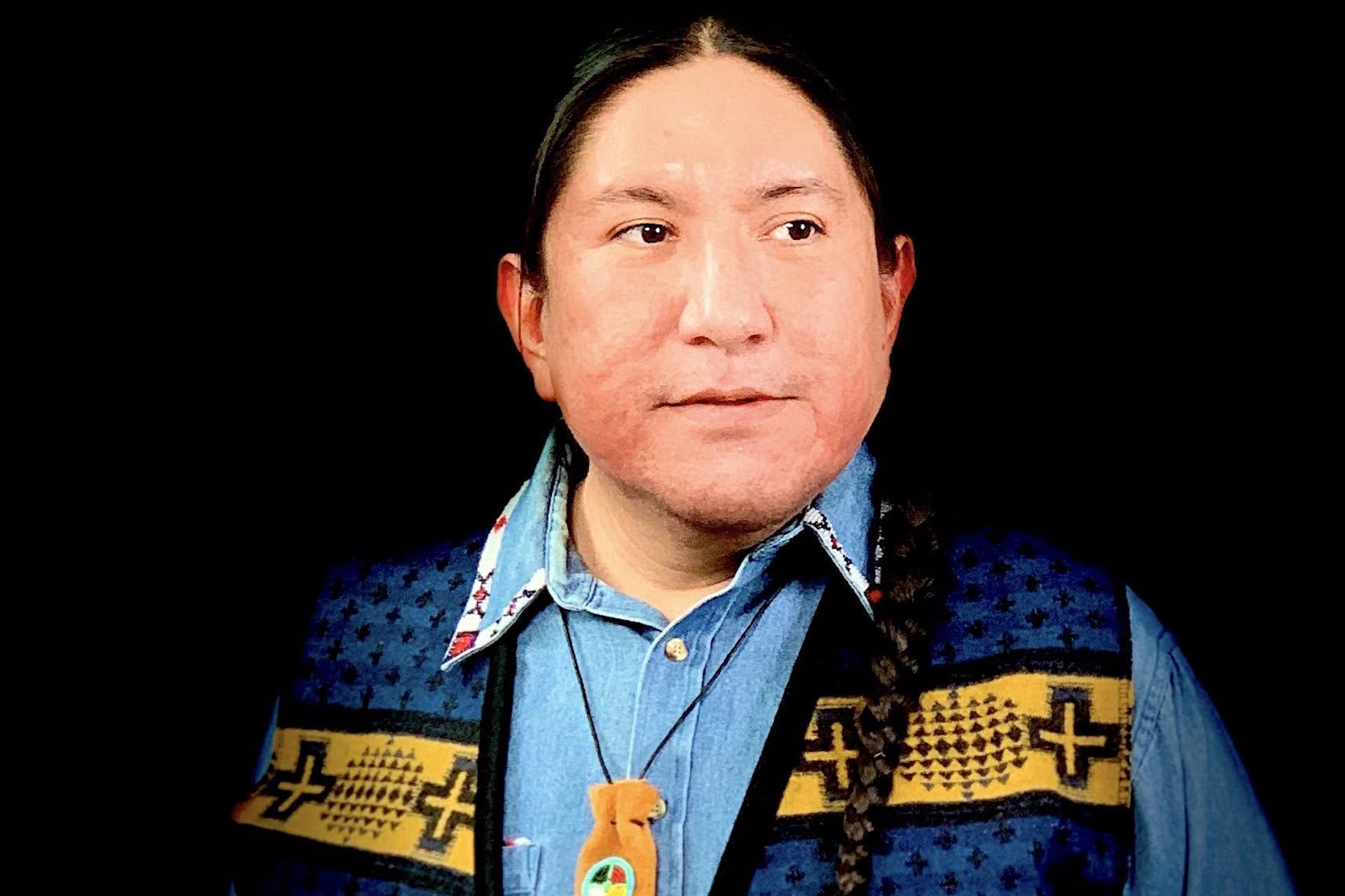 A head-and-shoulder shot of Michael Running Wolf. He is wearing a vest featuring Indigenous patterns and has a black braid that is draped over his left shoulder.