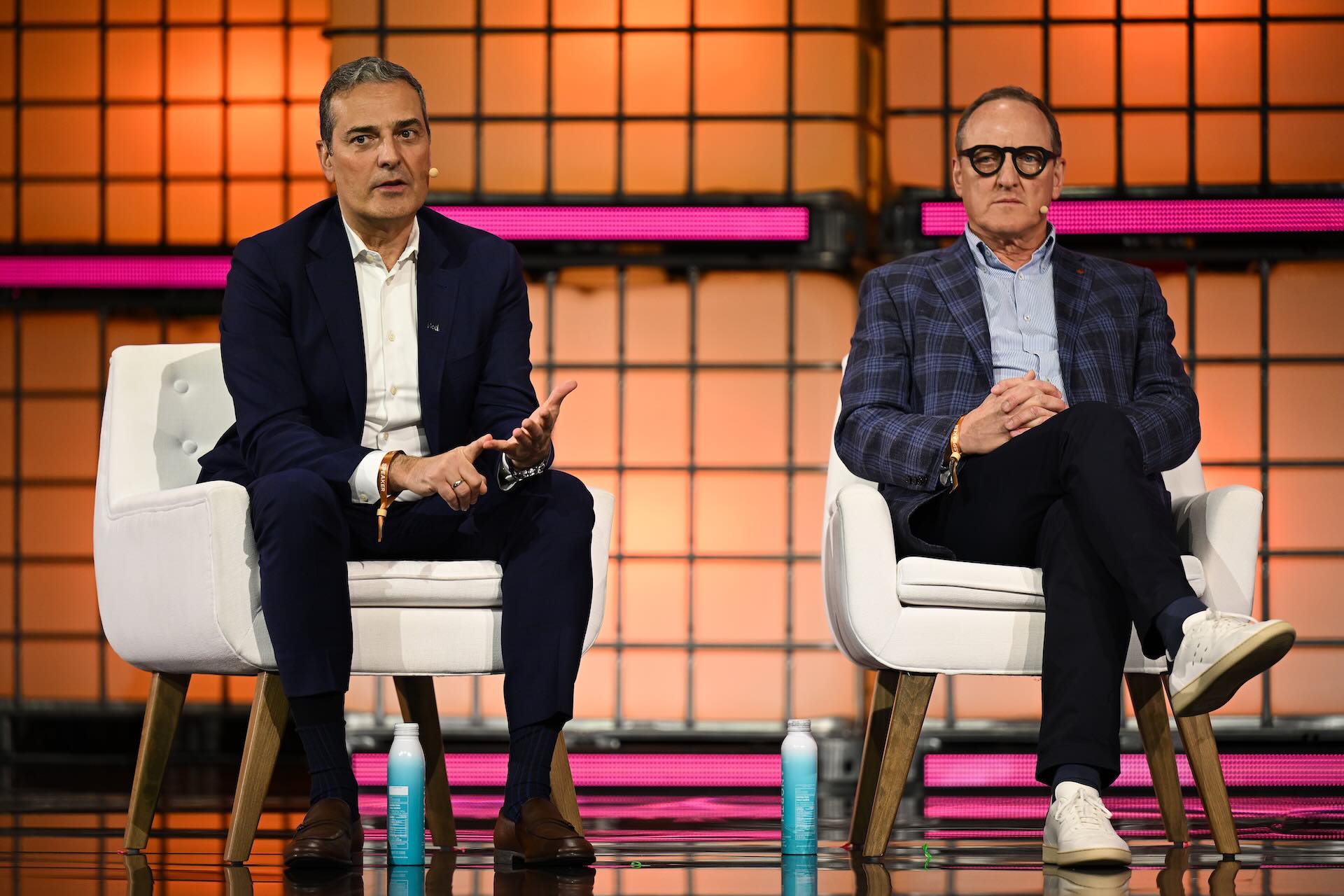 Bell CEO Mirko Bibic, left, and Coveo executive chair Louis Têtu, seated in white armchairs on a stage during a Web Summit Vancouver panel, with a pink-lit backdrop of latticed screens behind them.