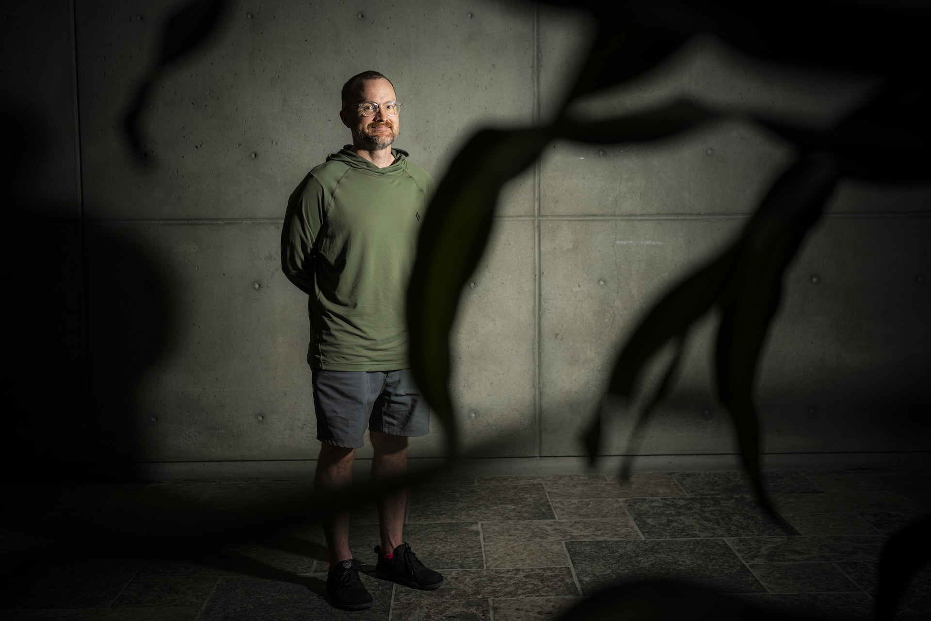 A portrait of Sam Witherspoon standing in front of an exposed concrete wall, taken from behind some plant fronds. He is wearing shorts and an olive-coloured runner's hoodie.