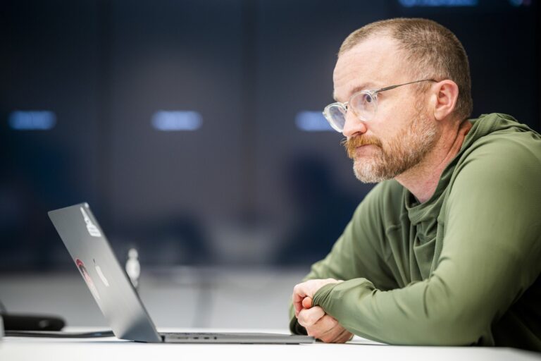 A shot of Sam Witherspoon in front of his laptop, looking pensively over the screen. He's wearing an olive-green hoodie.