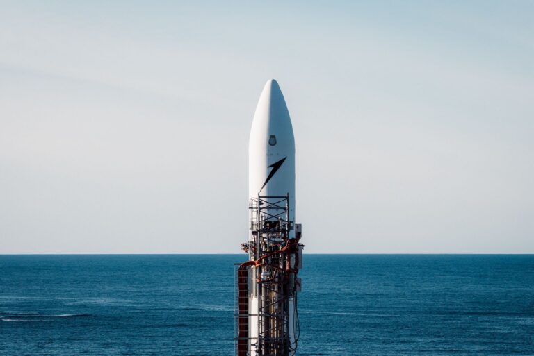 A shot of a small rocket sitting on a launch pad attached to its launch equipment. The backdrop is open sea and a light blue sky.