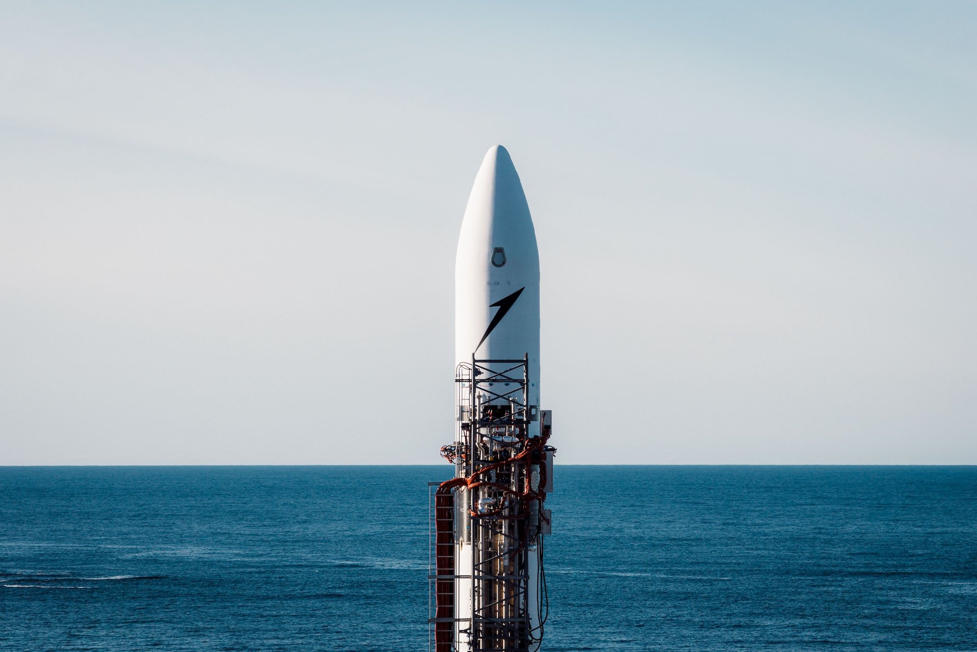 A shot of a small rocket sitting on a launch pad attached to its launch equipment. The backdrop is open sea and a light blue sky.