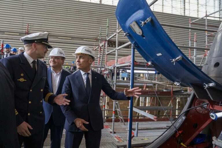 A shot of Mark Carney in a hardhat speaking to a German naval officer. They are standing in a small group on a scaffold deck, beside the open hatch of a submarine.