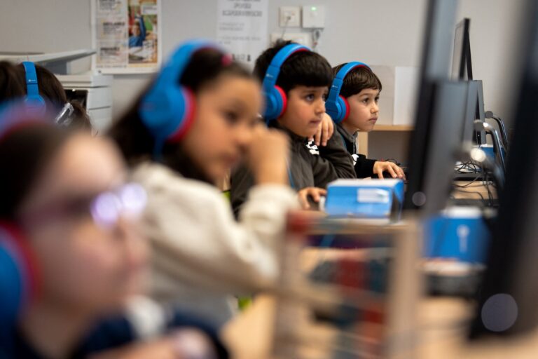 A row of school children at computers wearing brightly coloured headphones.