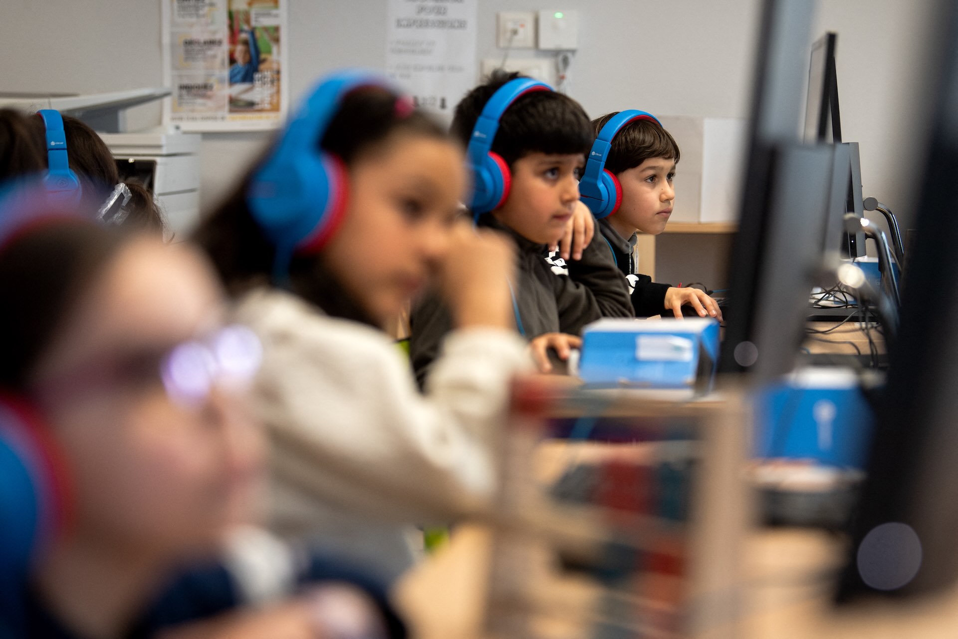 A row of school children at computers wearing brightly coloured headphones.