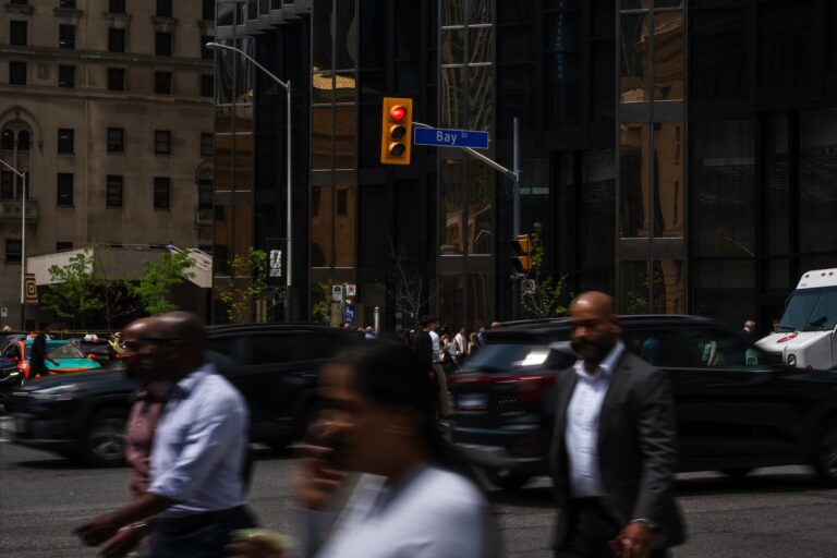 Three people cross a busy intersection on Bay Street, against a backdrop of dark buildings and cars. Their silhouettes are blurred.