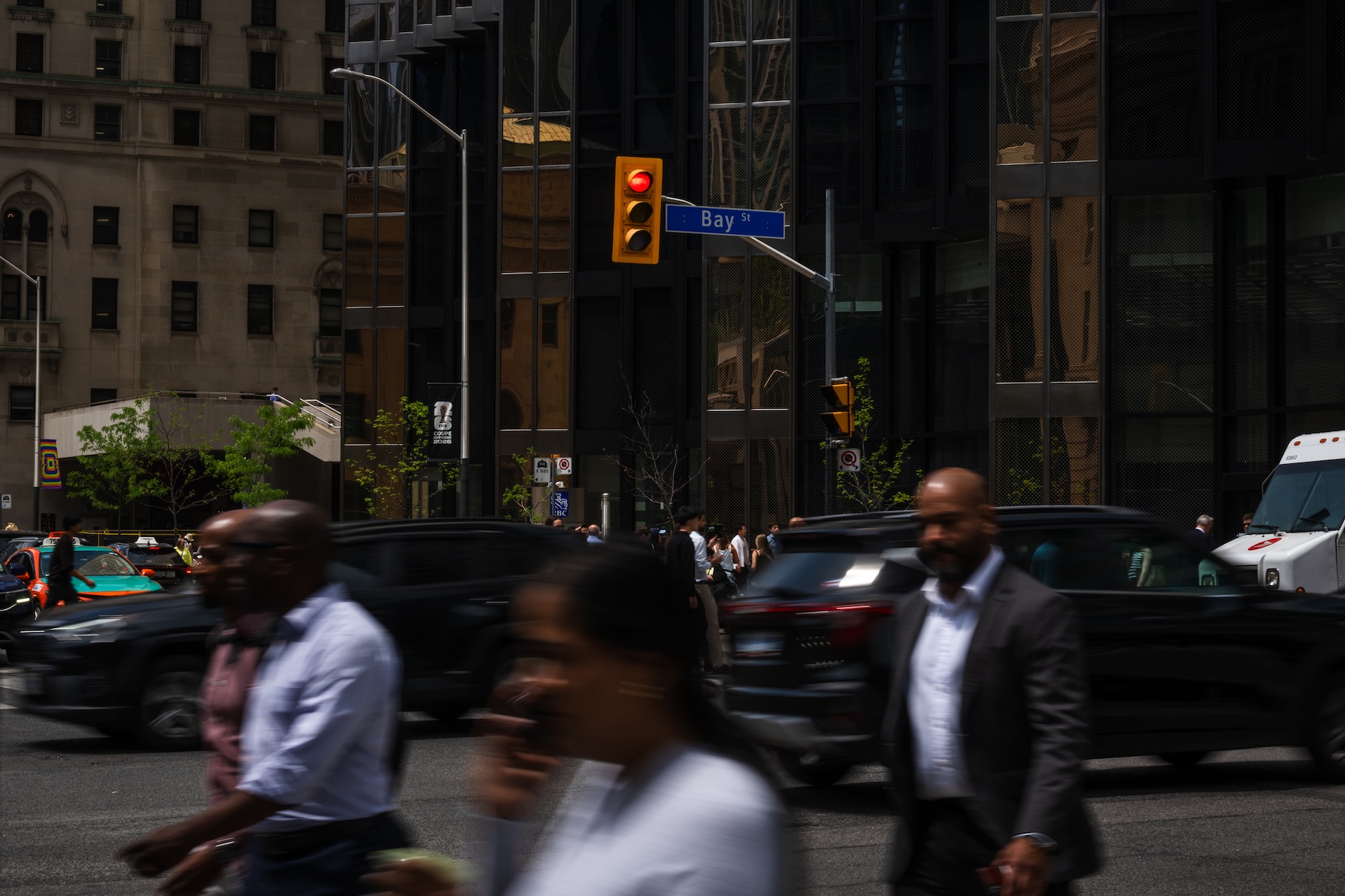 Three people cross a busy intersection on Bay Street, against a backdrop of dark buildings and cars. Their silhouettes are blurred.