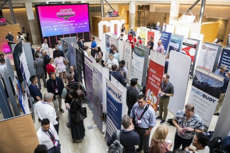 People stand in large room split into several aisles by banners promoting startups. A video screen on the back wall has the logo for the CDL Super Session on it.