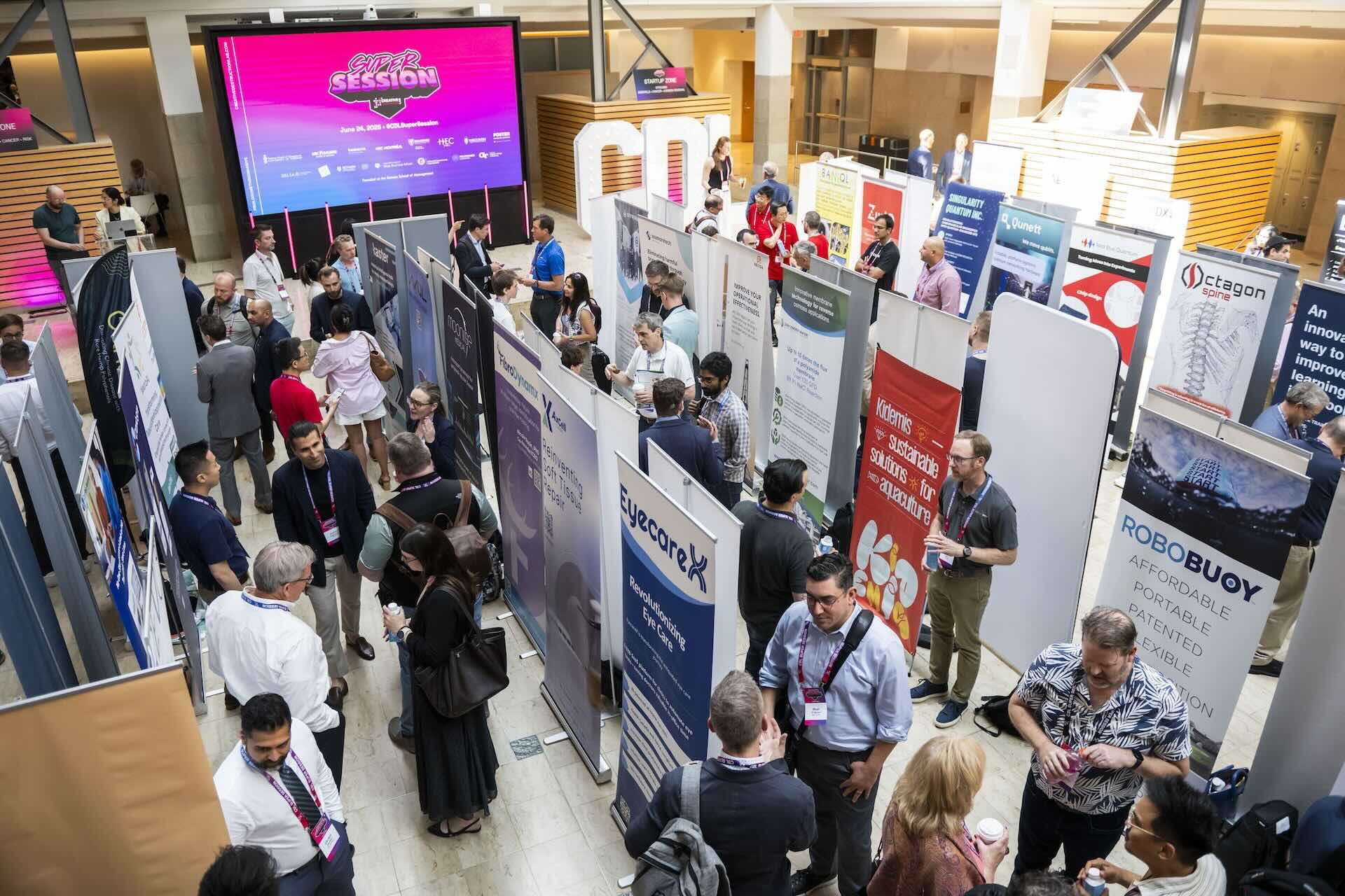 People stand in large room split into several aisles by banners promoting startups. A video screen on the back wall has the logo for the CDL Super Session on it.