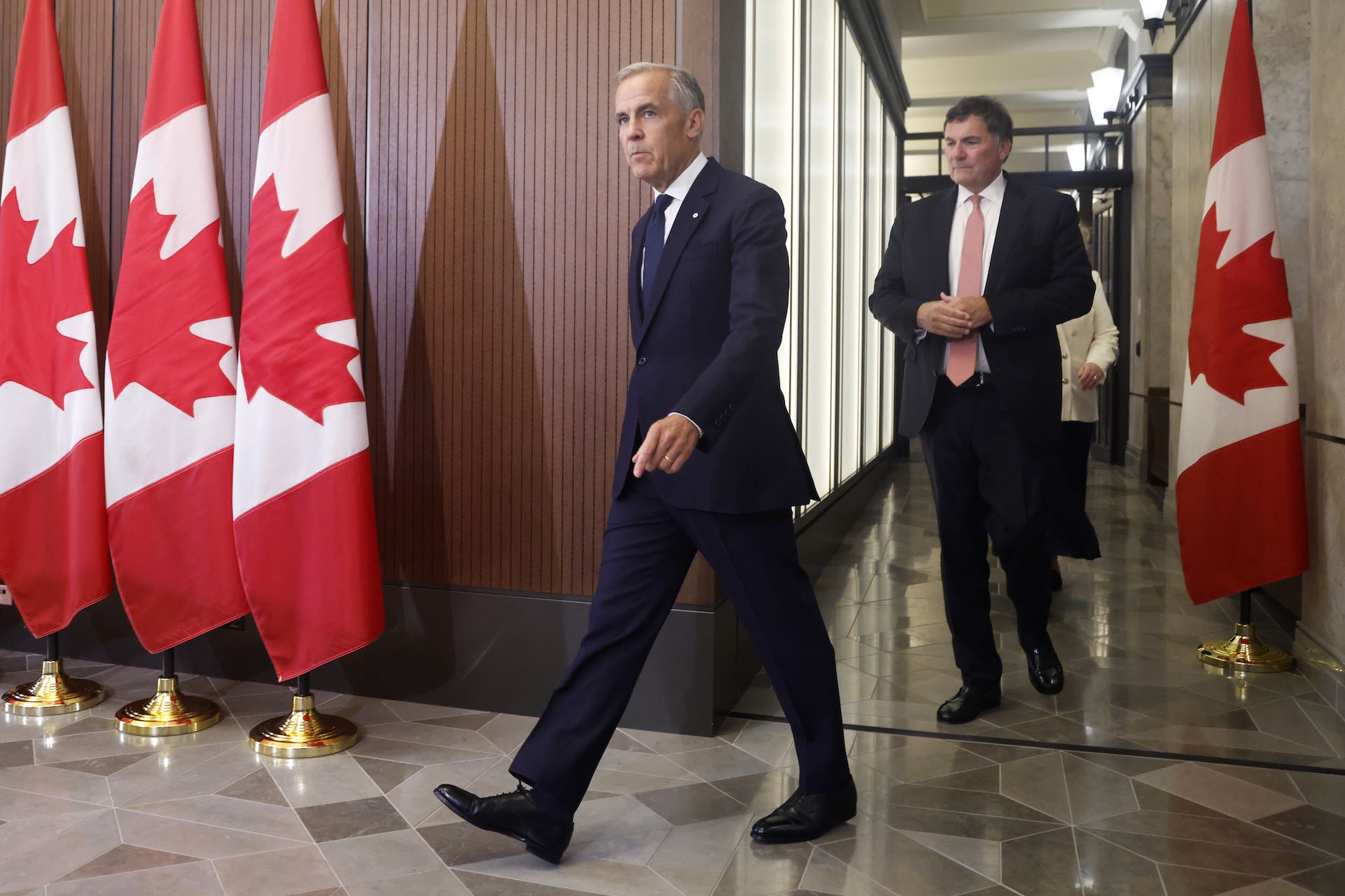 A photo of Mark Carney on his way into a news conference, trailed by Dominic LeBlanc. There is a row of Canadian flags on stands behind the lectern Carney is approaching.