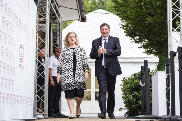 Janice Charette and Dominic LeBlanc walking toward the camera next to a white pavilion that bears the logo of the 2026 G7 meeting in France.