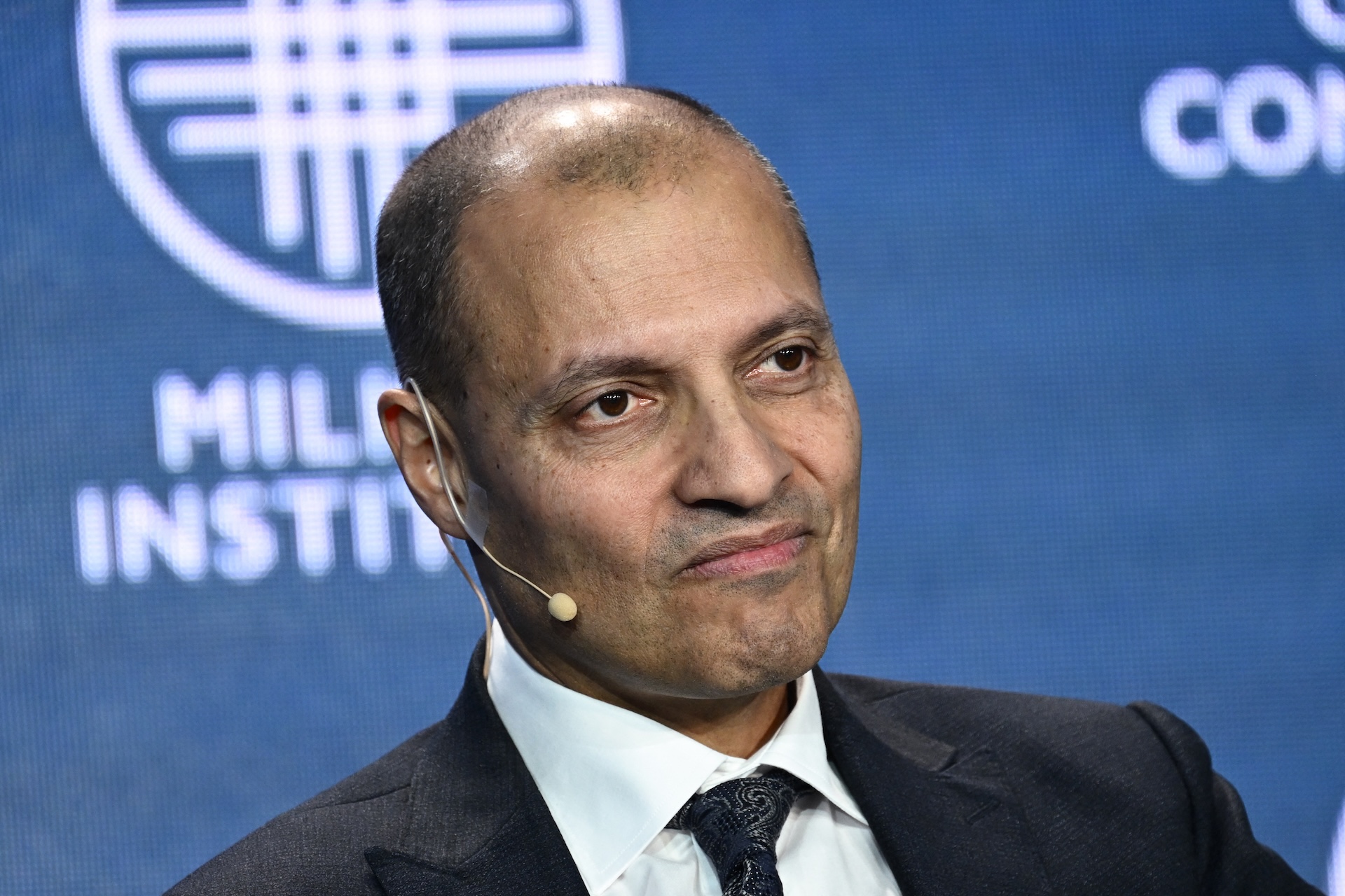 An image of a man wearing a dark suit jacket and tie. He is wearing a mic and looking slightly away from the camera.