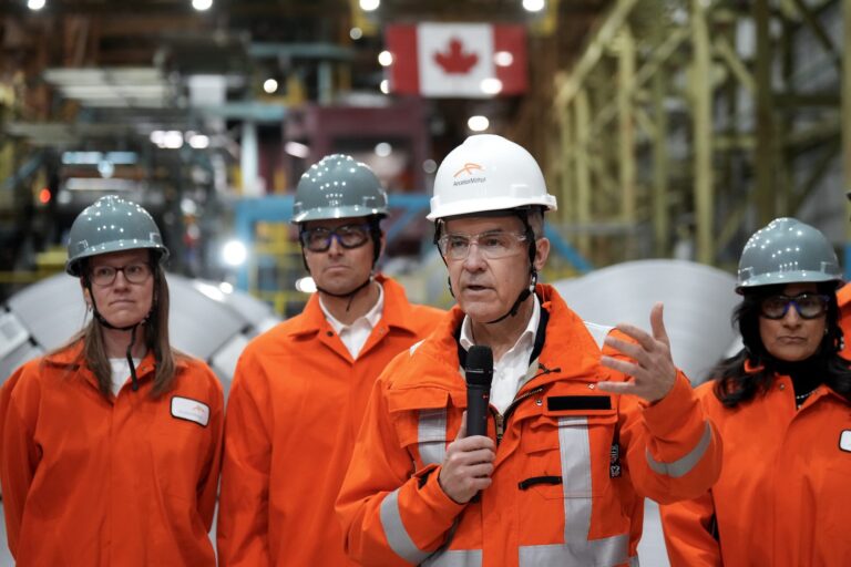 Four people stand in orange safety jackets and helmets stand in a factory, including Prime Minister Mark Carney, second right, speaking into microphone. They are in a plant with yellow scaffolding and a Canadian flag in the background.