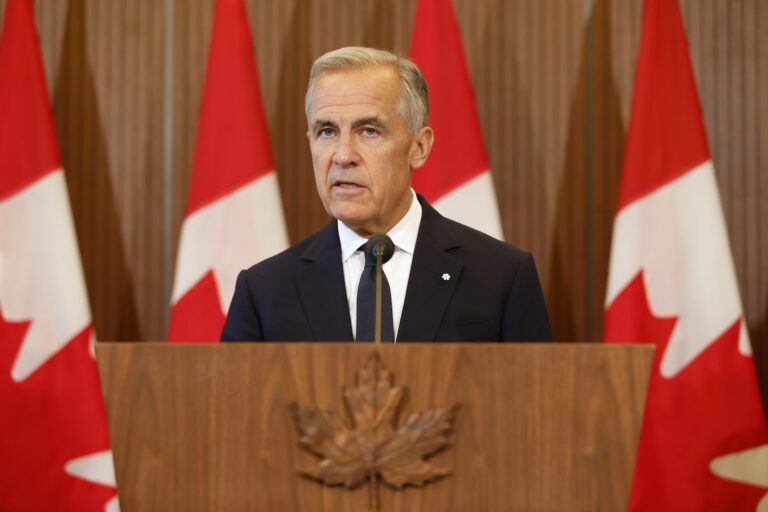 A head-on shot of Mark Carney at a lectern embossed with a maple leaf carved from wood. There is a row of Canadian flags behind him, and he's looking at the camera with a serious expression.