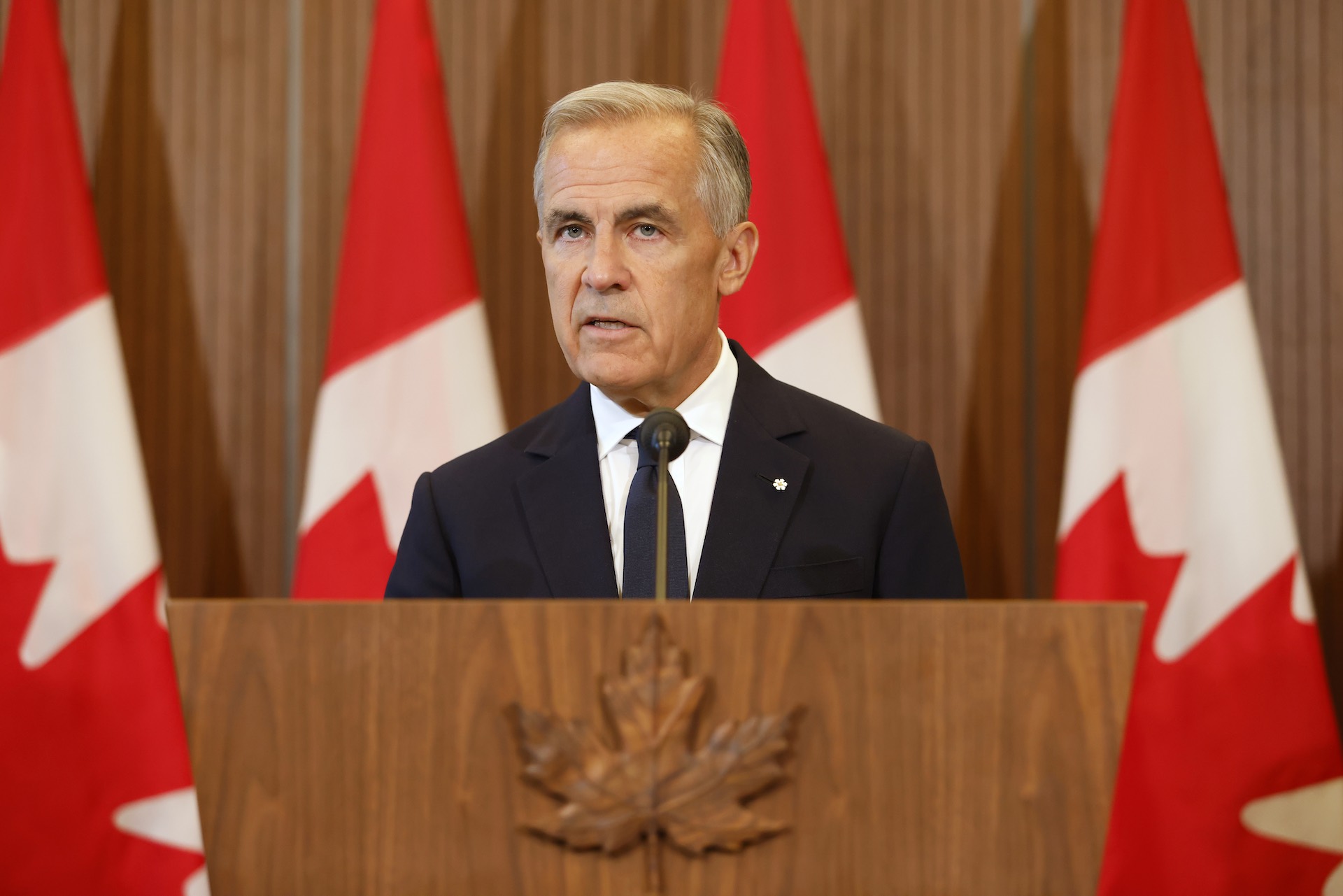 A head-on shot of Mark Carney at a lectern embossed with a maple leaf carved from wood. There is a row of Canadian flags behind him, and he's looking at the camera with a serious expression.