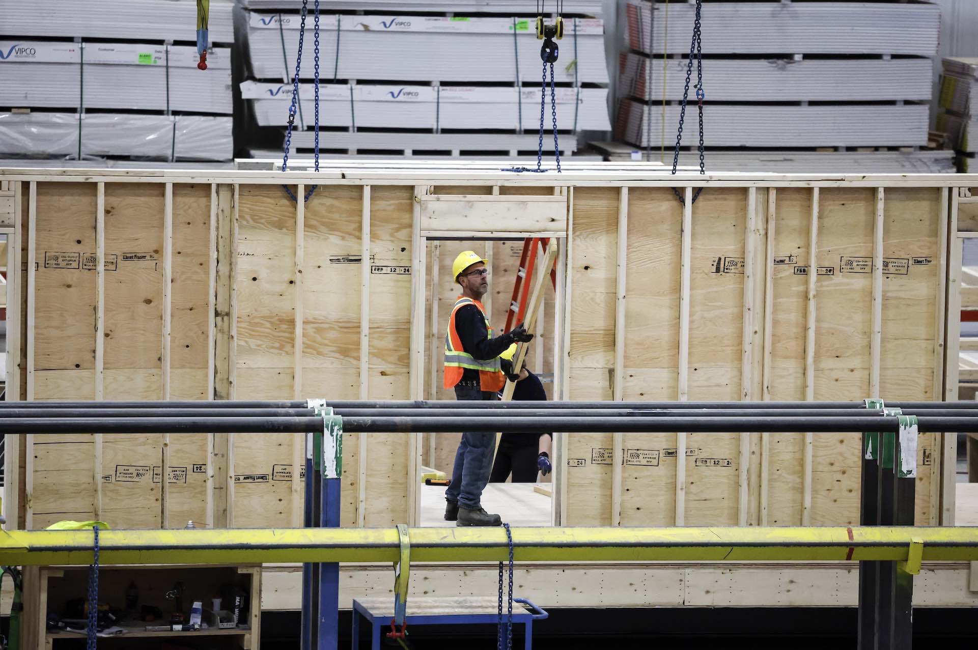 A shot of a construction worker seen through the door opening of a newly framed wall. The wall is being constructed in a factory setting.