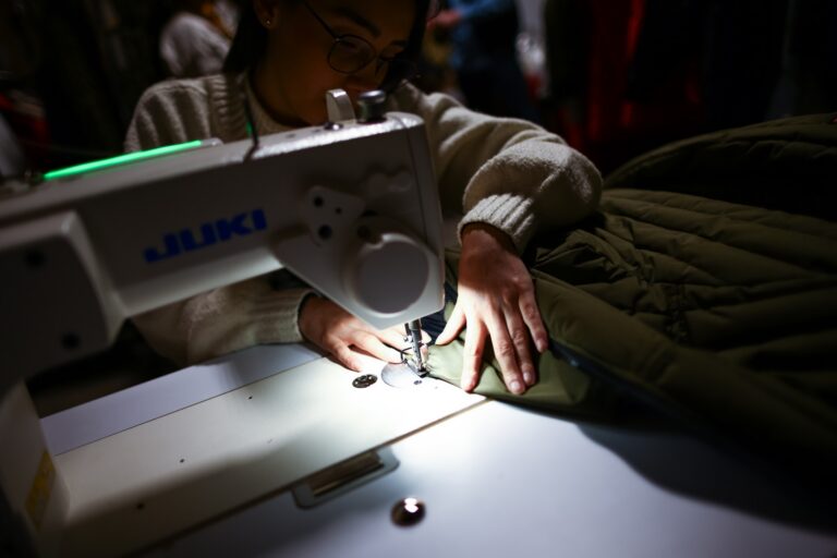 A close-up of a worker machine-sewing seams into an olive-green parka.