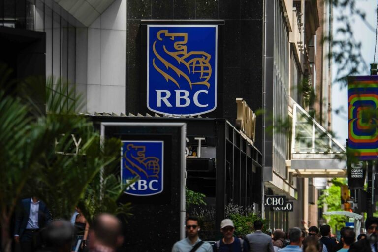 An RBC storefront sign with blue and gold lion-and-globe-logo, mounted on a dark building facade above a street in Toronto, as pedestrians pass by.