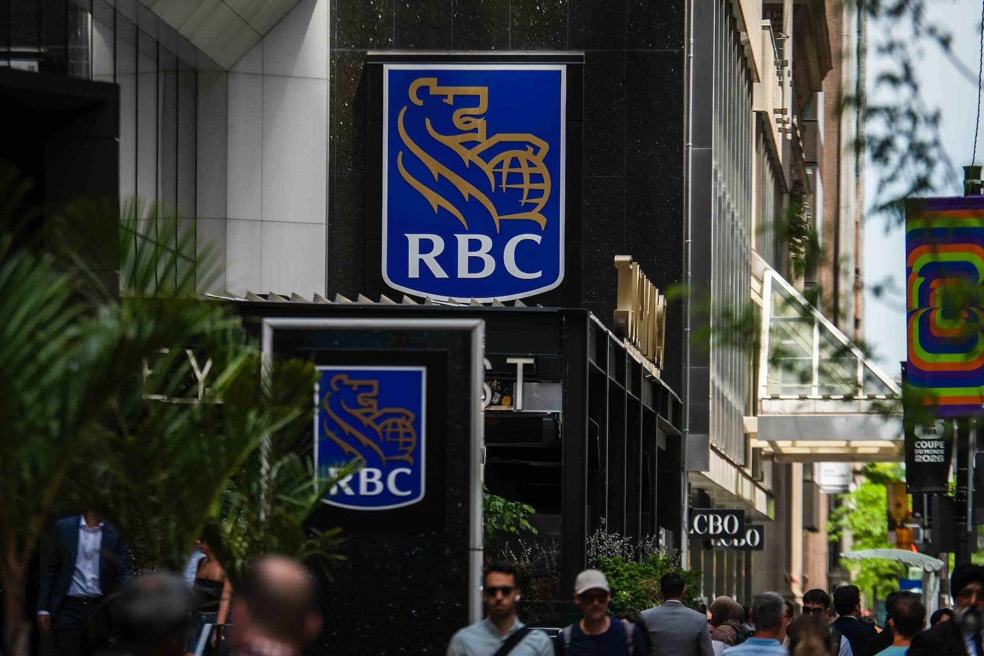 An RBC storefront sign with blue and gold lion-and-globe-logo, mounted on a dark building facade above a street in Toronto, as pedestrians pass by.
